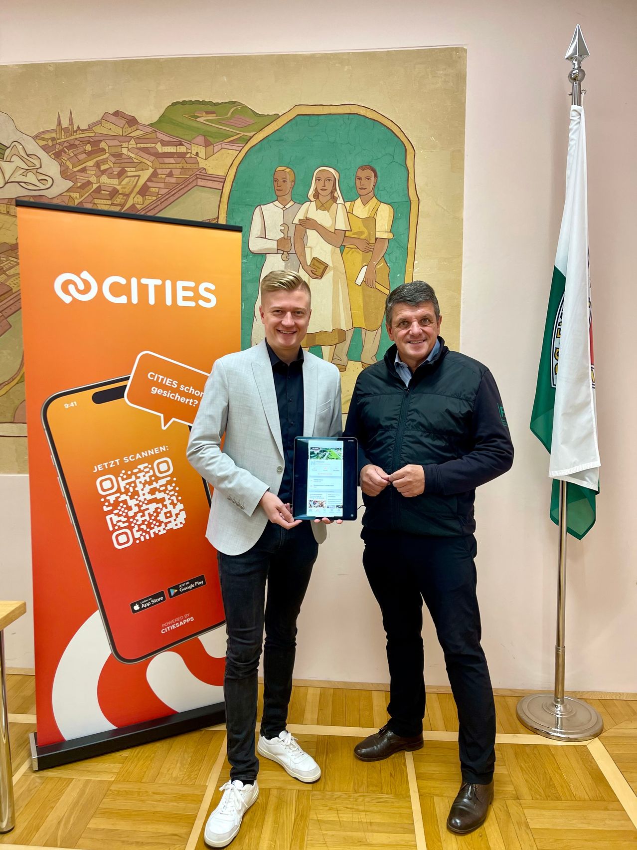 Zwei Männer stehen vor einem Banner mit einem QR-Code und halten ein Tablet. Dahinter befindet sich eine Flagge und eine Wandmalerei mit Menschen.