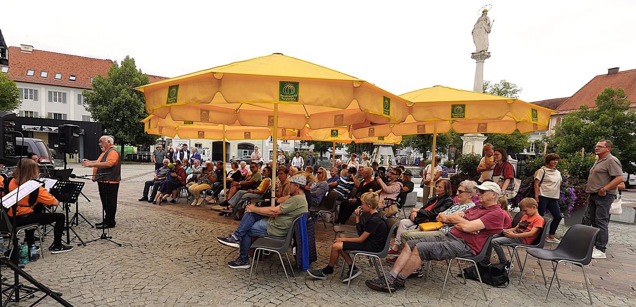 Eine Gruppe von Menschen sitzt unter gelben Schirmen in einer Außenumgebung. Sie schauen in Richtung einer Bühne oder eines Aufführungsbereichs. Im Hintergrund befinden sich Bäume und Gebäude.