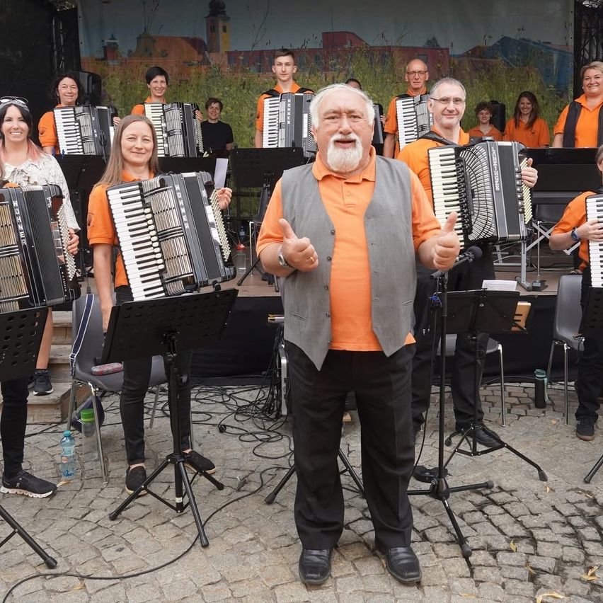 Eine Gruppe von Musikern tritt auf einer Bühne auf. Ein älterer Mann steht vor der Gruppe und lächelt. Sie tragen alle orangefarbene Shirts und halten Akkordeons.