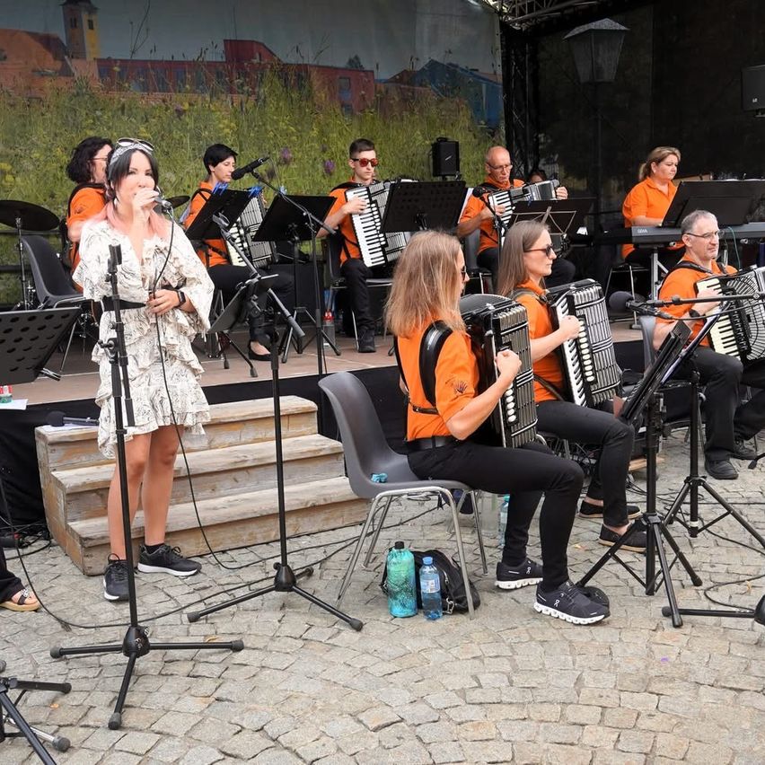 Eine Band tritt auf der Bühne auf. Die Frau singt, während andere Instrumente spielen, darunter Akkordeons. Es gibt Stühle, Mikrofone und Flaschen auf dem Boden.