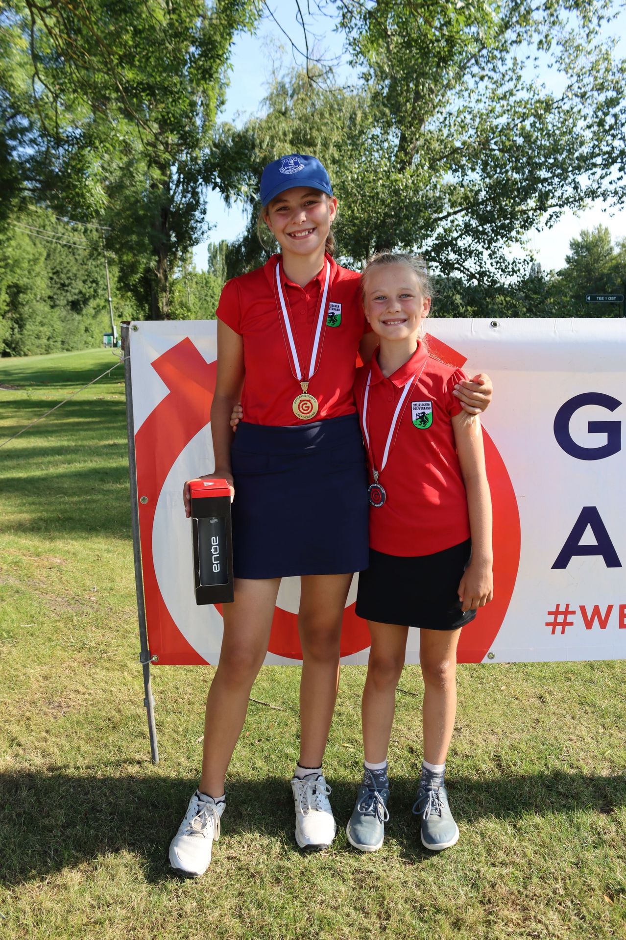 Zwei Mädchen stehen nebeneinander, beide tragen rote Shirts und Shorts. Sie haben Medaillen um den Hals. Das Mädchen auf der linken Seite hält eine Trophäe. Sie stehen vor einem Banner mit dem Text 'GA' und dem Hashtag '#WE'. Dahinter befindet sich eine Rasenfläche mit Bäumen.