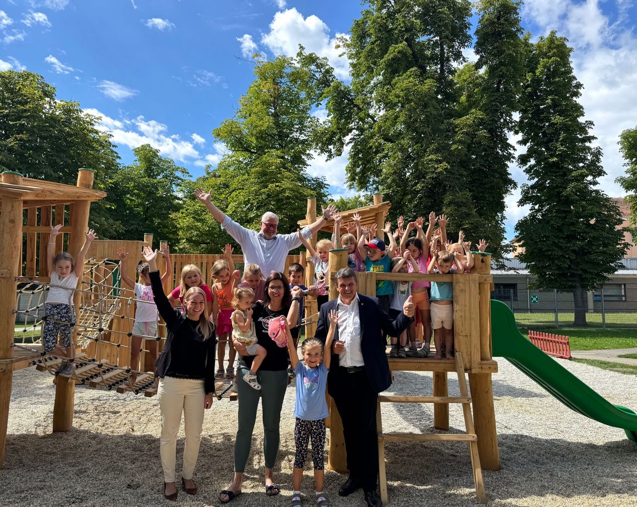 Eine Gruppe von Menschen, darunter Kinder, posiert für ein Foto auf einem Spielplatz mit einem Mann im Anzug und zwei Frauen vor ihnen.