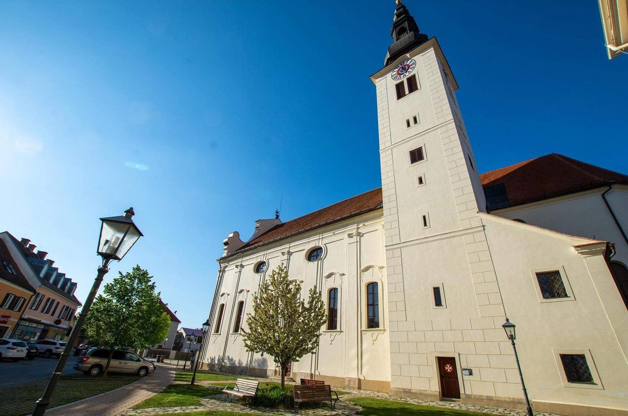 Eine weiße Kirche mit einem hohen Turm, einer Uhr und einem Kreuz auf der Spitze. Die Kirche hat mehrere Bogenfenster und ein braunes Dach. Vor der Kirche stehen zwei Bänke auf dem Rasen.