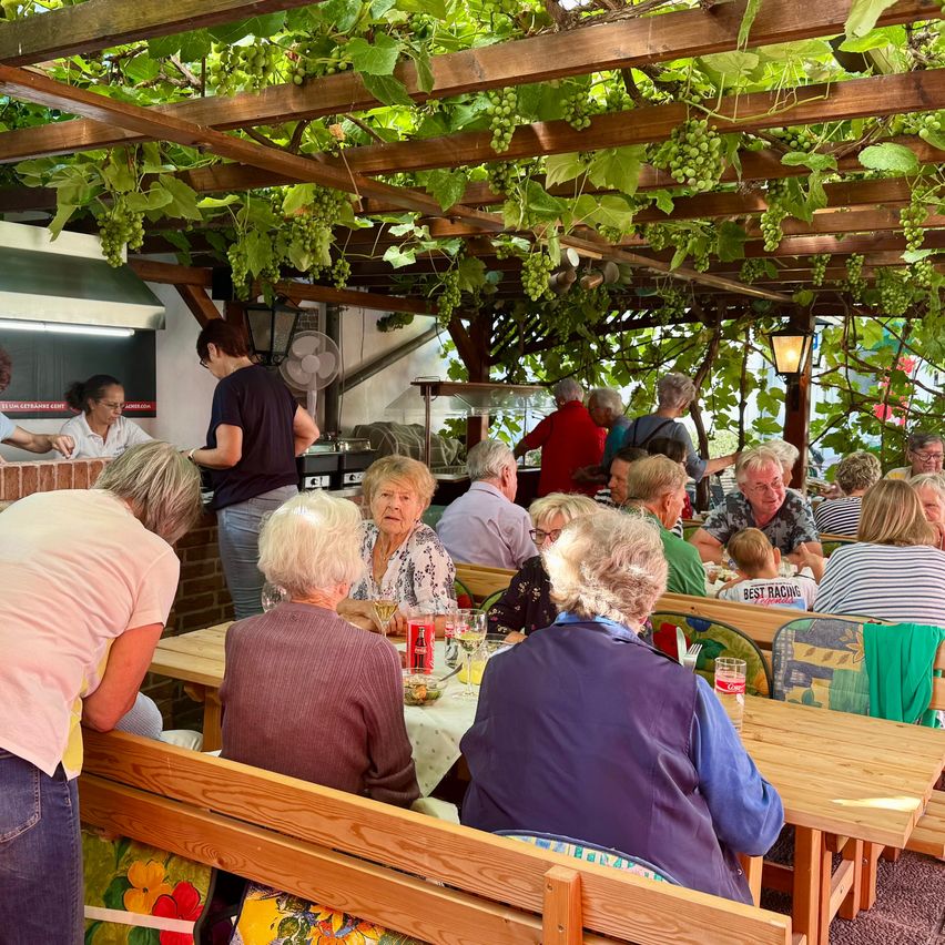 Eine Gruppe älterer Menschen sitzt an einem Tisch in einem Restaurant im Freien. Eine Person steht hinter der Theke und eine Lampe hängt von der Decke.