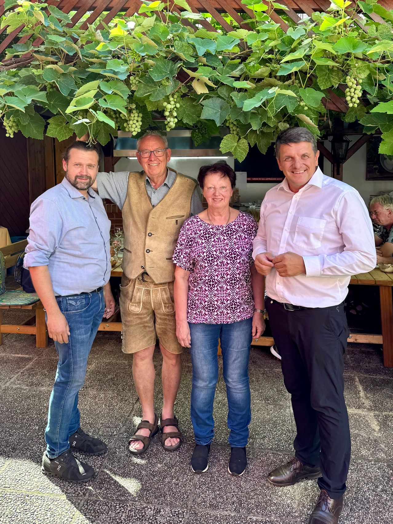 Vier Personen posieren für ein Foto unter einem Weinlaub. Der Mann links trägt ein blaues Hemd und Jeans. Der zentrale Mann trägt eine braune Weste und Shorts. Die Frau in der Mitte trägt ein gemustertes Oberteil und Jeans. Der Mann rechts trägt ein weißes Hemd und dunkle Hosen. Hinter ihnen ist ein Holztisch zu sehen.