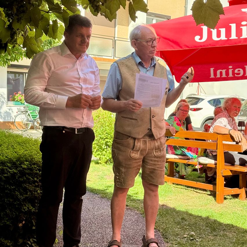 Zwei Männer stehen unter einem roten Regenschirm, einer hält ein Stück Papier und gestikuliert, während der andere zuhört. In der Nähe sitzt eine Holzbank mit Menschen. Ein Fahrrad ist geparkt und ein Auto ist im Hintergrund.