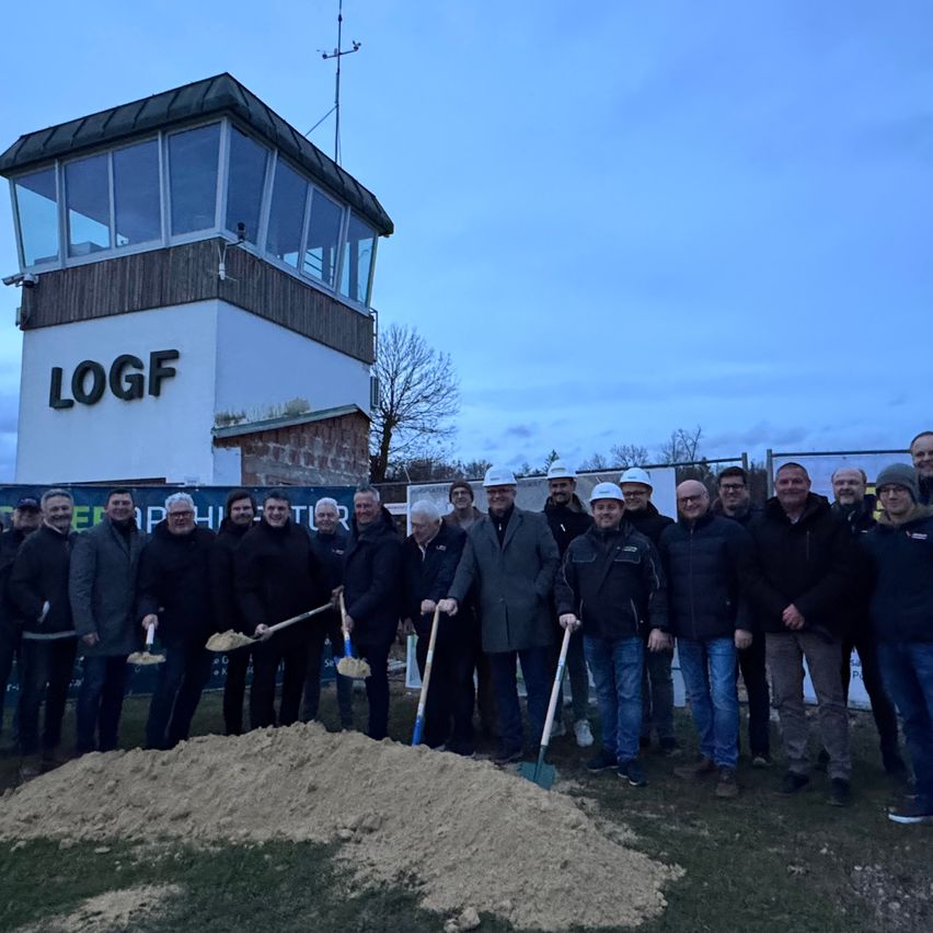 Eine Gruppe von Menschen posiert für ein Foto vor einem LOGF-Turm. Sie halten Schaufeln in der Hand und stehen auf einem Haufen Erde.