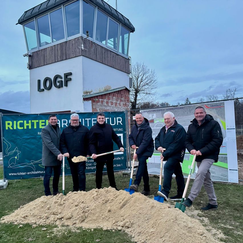 Fünf Männer mit Schaufeln stehen lächelnd vor einem Haufen Erde. Dahinter befindet sich ein Banner mit 'RICHTER ARCHITEKTUR' und ein Gebäude mit 'LOGF'. Links steht ein Turm mit einem Glasfenster.