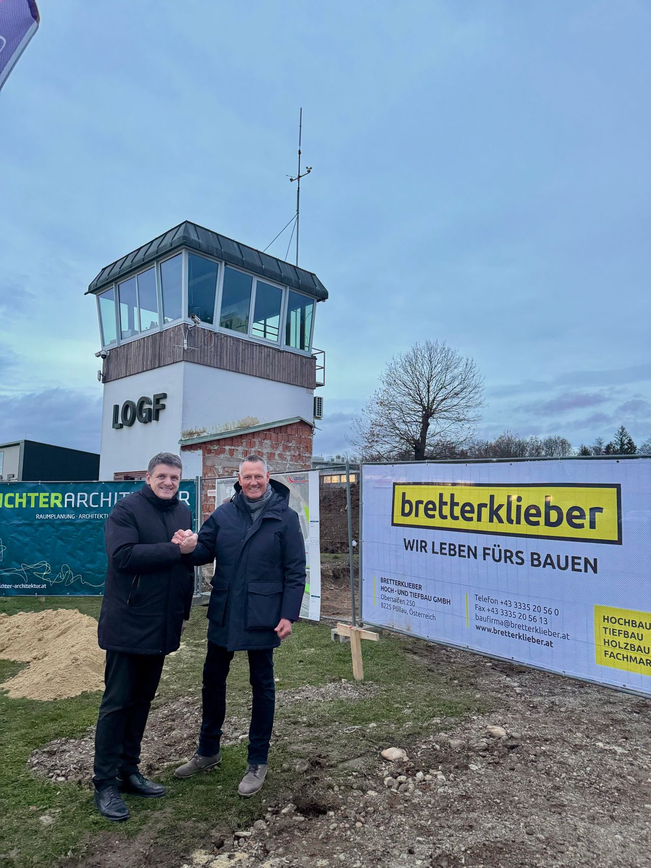Zwei Männer stehen vor einem Gebäude mit einem Turm, auf dem LOGF steht. Sie lächeln und scheinen sich die Hände zu schütteln. Hinter ihnen befindet sich ein Banner, das 'bettlerklieber' bewirbt.