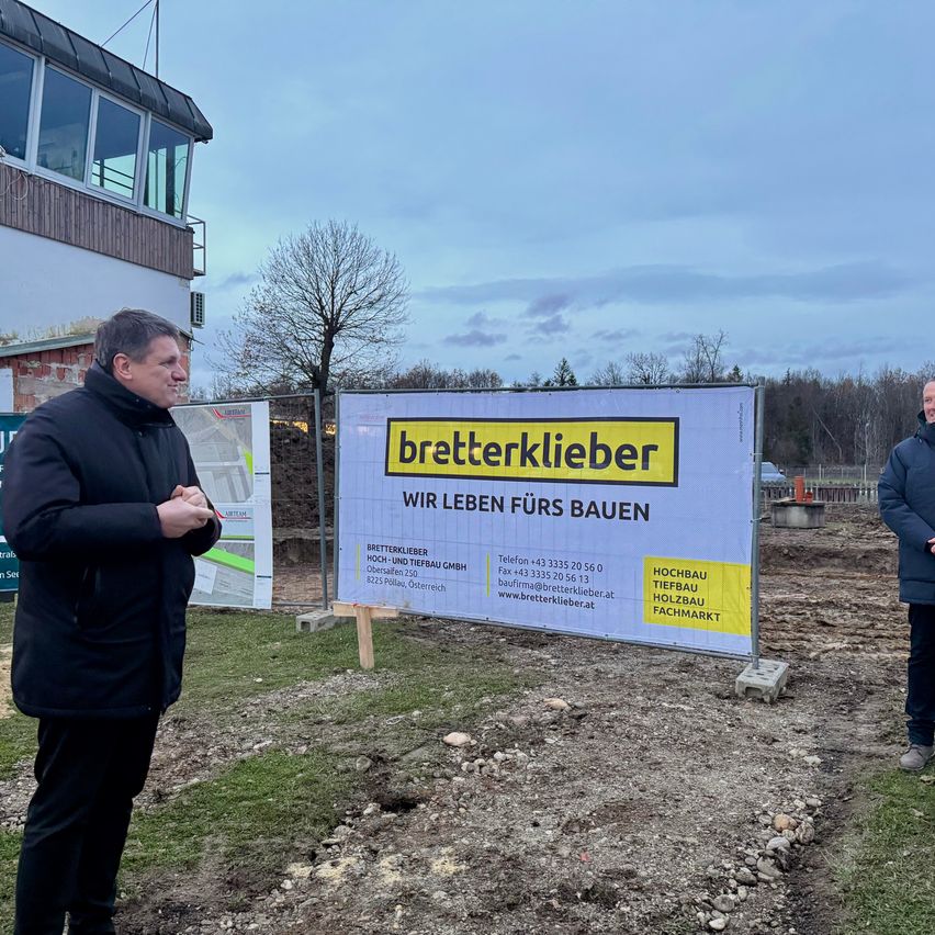 Zwei Männer stehen vor einem Gebäude im Bau, mit einem großen Schild hinter ihnen. Das Schild trägt die Aufschrift 'bretterklieber' mit dem Slogan 'Wir leben fürs Bauen'. Ein Mann hält einen Stift und scheint zu sprechen.
