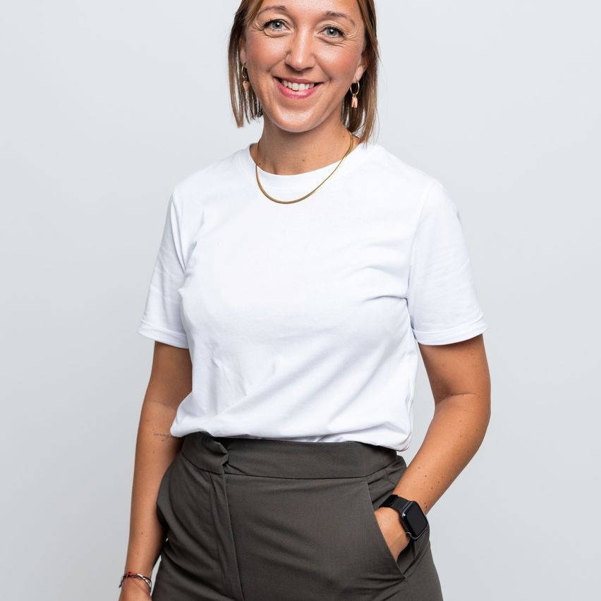 Eine Frau in einem weißen T-Shirt und grauer Hose lächelt und posiert für ein Foto mit einer Hand in der Tasche.