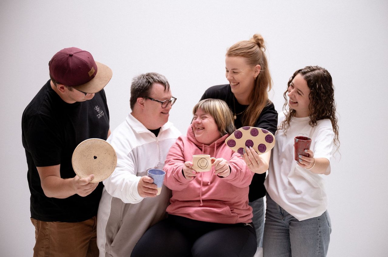 Fünf Menschen lächeln und lachen und halten verschiedene Keramikgegenstände. Eine Frau in einer rosa Kapuzenjacke hält einen Becher mit einem lächelnden Gesicht. Ein Mann mit Brille hält einen blauen Becher. Eine andere Frau hält einen keksförmigen Teller. Ein Mann mit einer Kappe hält einen runden Teller.