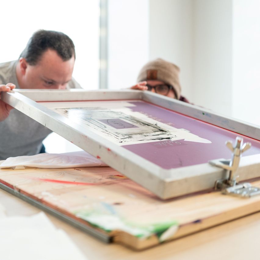 Zwei Personen arbeiten gemeinsam an einem Siebdruckprojekt. Ein Mann untersucht den Bildschirm, während der andere beobachtet. Der Bildschirm zeigt einen lila Hintergrund mit schwarzen und weißen Designs.