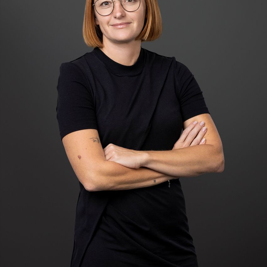 Eine Frau mit kurzen roten Haaren und Brille steht mit verschränkten Armen und lächelt direkt in die Kamera. Sie trägt ein schwarzes T-Shirt und hat Tätowierungen auf ihrem linken Arm. Der Hintergrund ist dunkel.