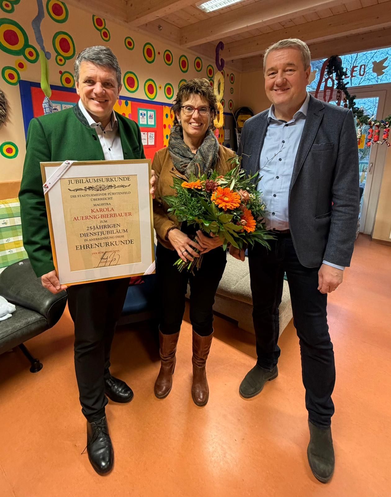 Drei Personen stehen in einem Raum, zwei Männer und eine Frau. Die Frau hält einen Blumenstrauß, und der Mann links hält ein Zertifikat. Der Mann rechts lächelt. Hinter ihnen ist eine Wand mit bunten Dekorationen.