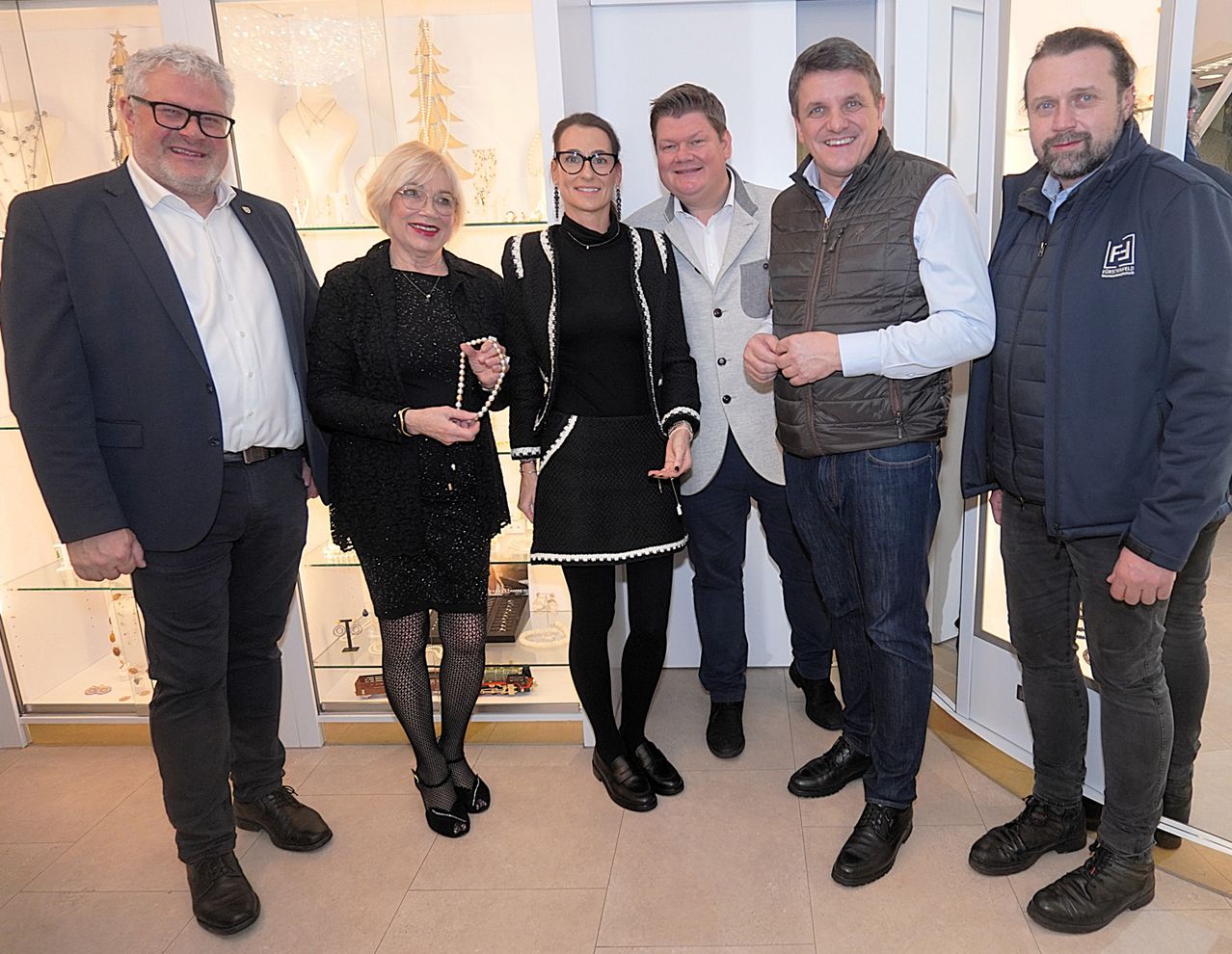 Fünf Personen stehen lächelnd für ein Foto in einem Raum. Eine Frau in Schwarz hält eine Perlenkette. Sie stehen vor einer Glasvitrine mit Schmuck.