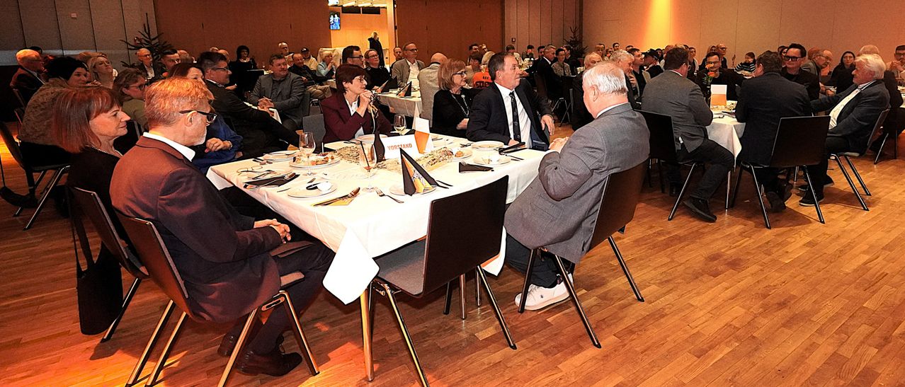 Mehrere Personen sitzen in einem Konferenzsaal an einem Tisch mit Tellern, Weingläsern und Servietten. Eine Person steht. Ein Mann mit Krawatte schaut einen anderen Mann an, der mit verschränkten Händen sitzt.
