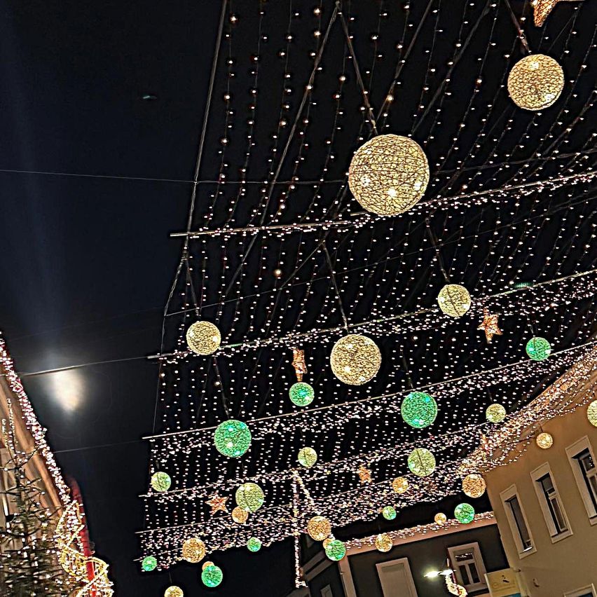 Eine Straße ist mit verschiedenen farbigen Lichtern dekoriert, darunter gelbe und grüne Kugeln, Sterne und Lichterketten. Die Gebäude auf beiden Seiten der Straße haben ebenfalls Weihnachtsdekorationen.
