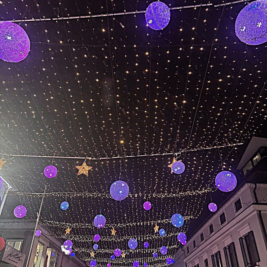 Ein nächtlicher Straßenblick, geschmückt mit lila und blauen Lichtern, Sternen und hängenden Ornamenten. Der Himmel ist dunkel mit verstreuten Sternen, und Gebäude säumen die Seiten.