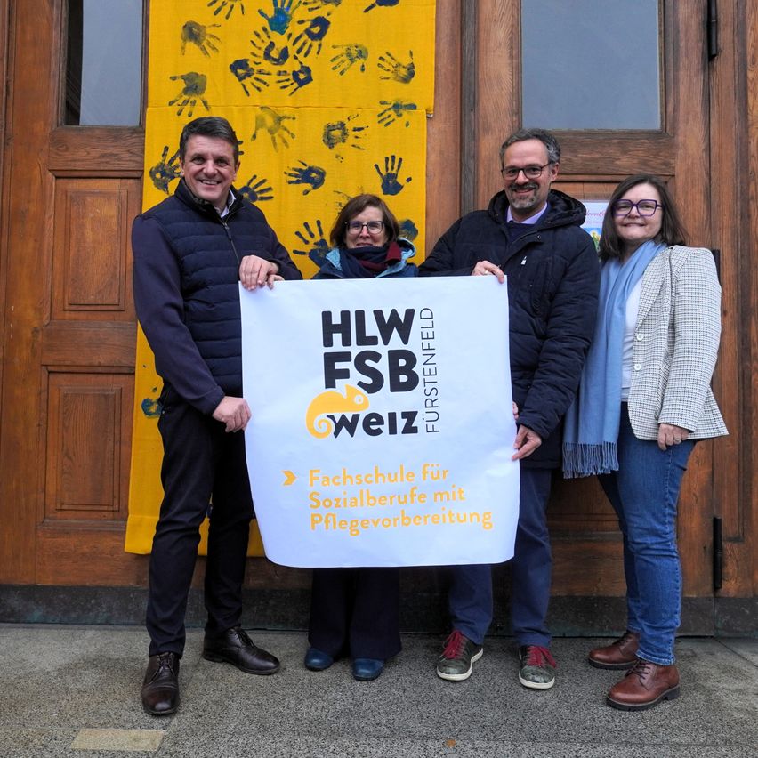 Vier Personen stehen vor einer Holztür und halten ein Banner mit der Aufschrift 'HLW FSB Weiz'. Sie lächeln und scheinen für ein Foto zu posieren. Hinter ihnen ist ein Banner mit Handabdrücken an der Wand befestigt.