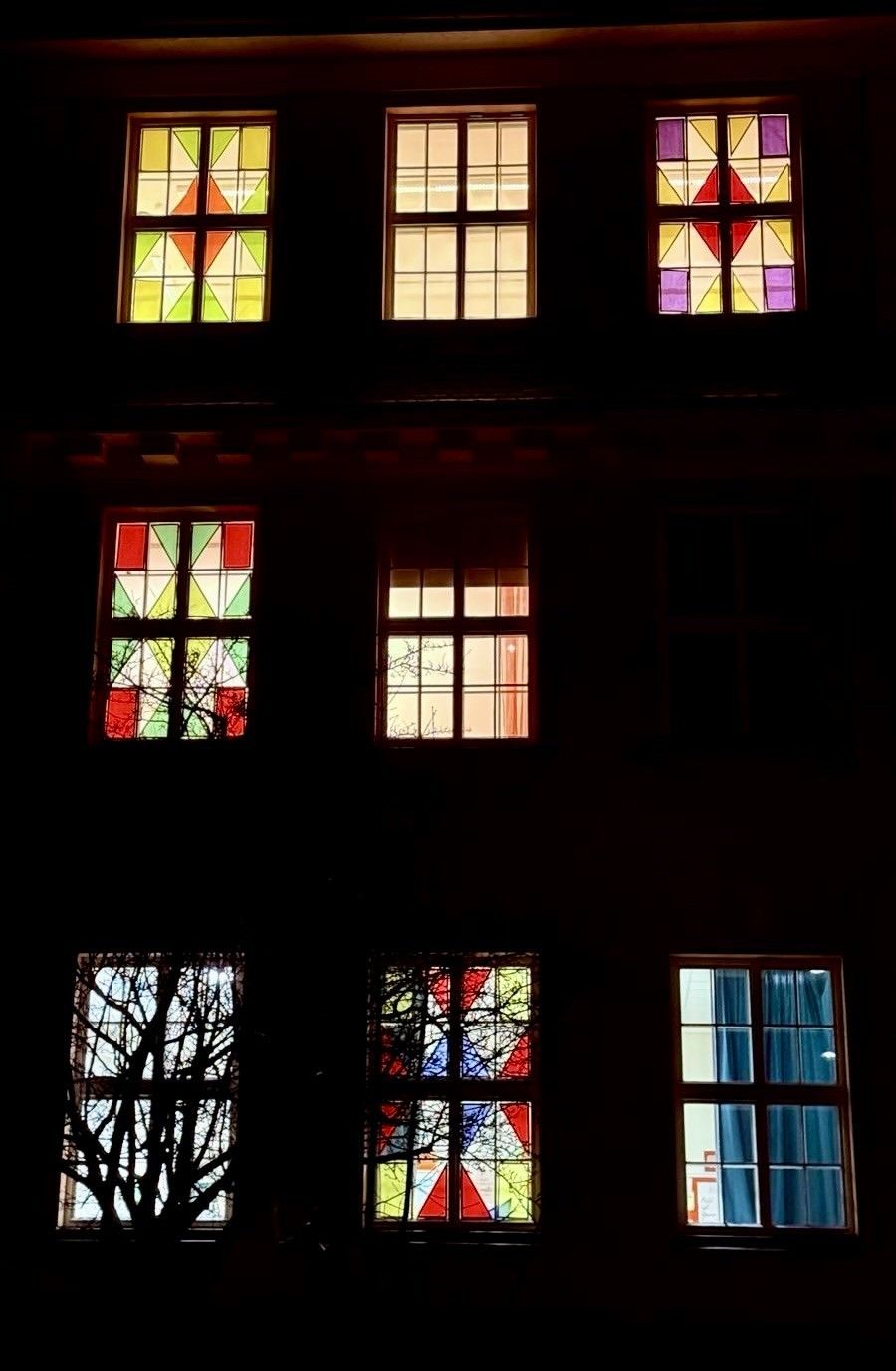 Ein Gebäude mit mehreren Buntglasfenstern ist von innen beleuchtet und wirft farbige Reflexionen. Die Fenster variieren in Design und Farbe.