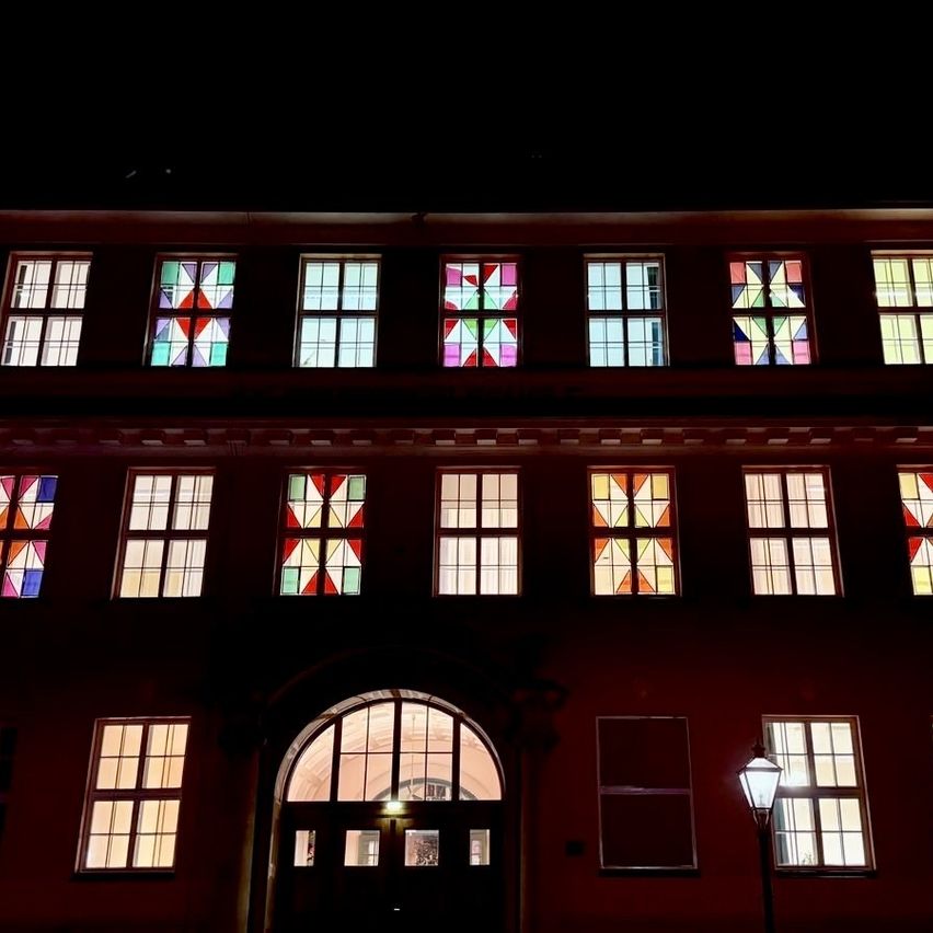Das Gebäude, nachts beleuchtet, zeigt eine Reihe von Fenstern mit farbenfrohen Buntglasdesigns. Das zentrale Bogenfenster und die Tür sind beleuchtet, mit einer Straßenlaterne rechts.