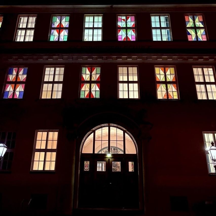 Das Gebäude ist nachts beleuchtet. Es hat viele Fenster, jedes mit einem farbenfrohen rautenförmigen Design. Der Haupteingang ist eine gewölbte Tür mit einem Fenster darüber. Es gibt zwei Lampen auf beiden Seiten des Eingangs.