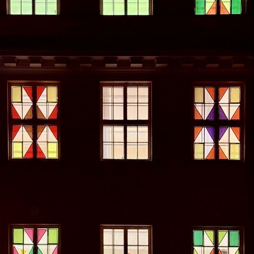 Ein Nahaufnahme der Fassade eines Gebäudes zeigt mehrere Fenster. Jedes Fenster hat ein einzigartiges Design, einige mit Buntglas und andere klar. Die Fensterrahmen sind dunkel und kontrastieren mit den hellen Farben des Glases.