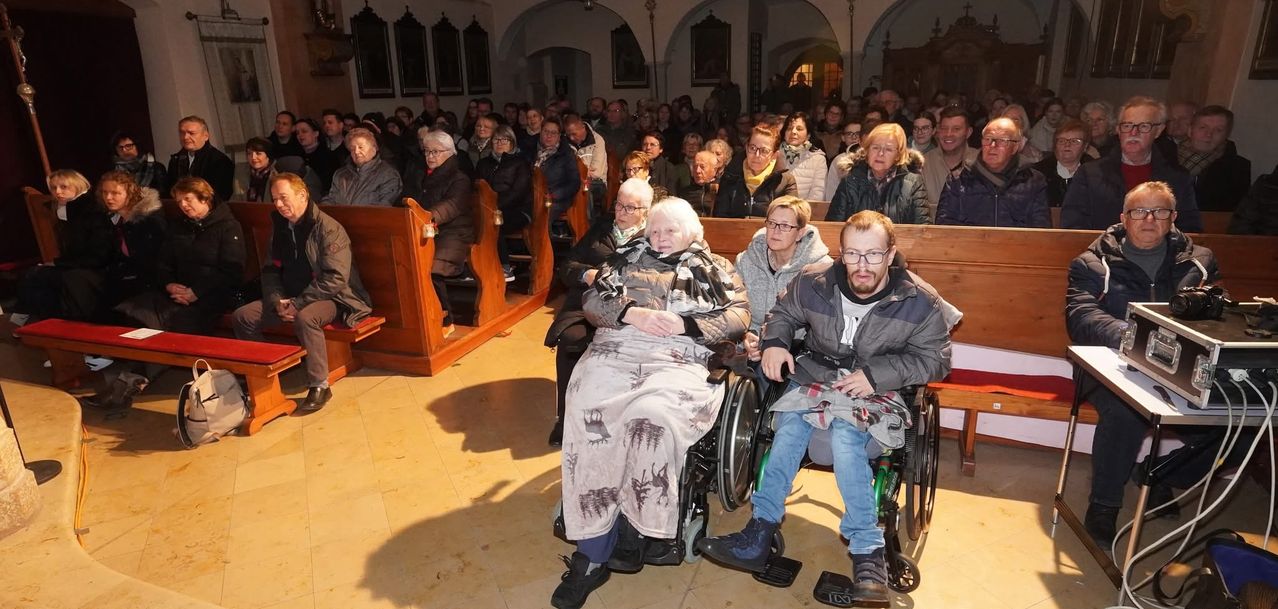 Eine Gruppe von Menschen in einer Kirche, einige in Rollstühlen, sitzen auf Kirchenbänken und schauen nach vorne. Viele tragen Brillen und Mäntel.