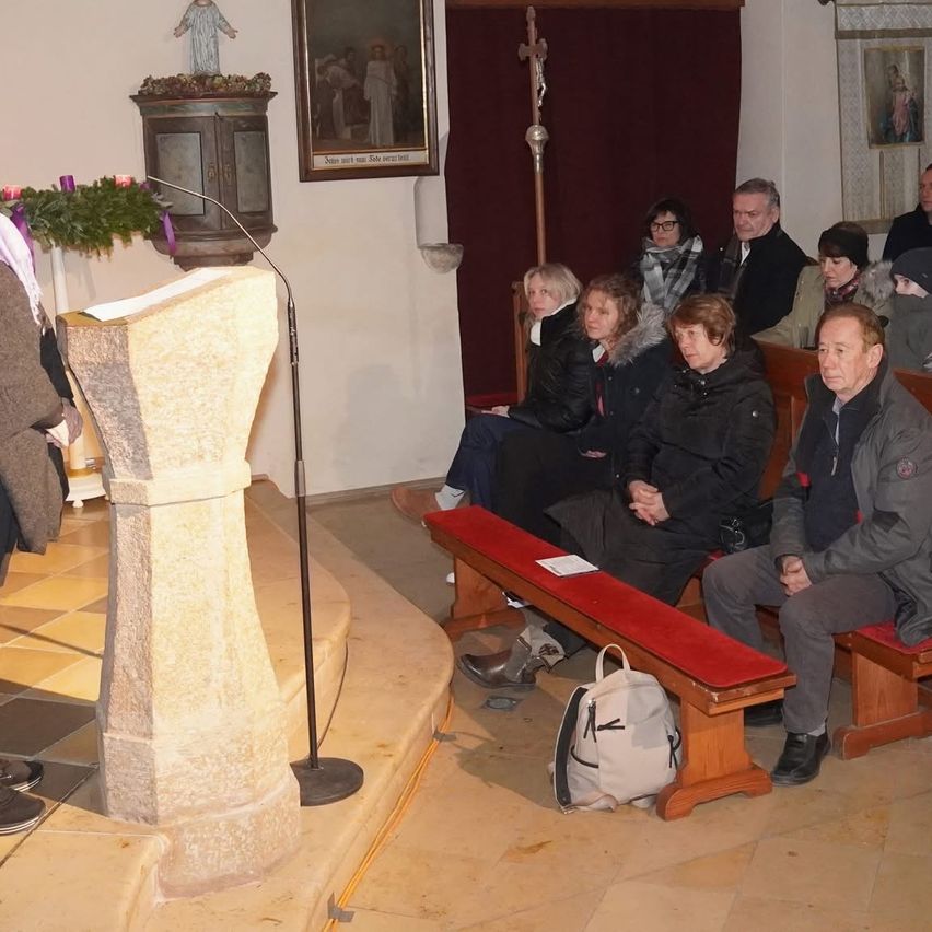 Mehrere Menschen sitzen auf Bänken in einer Kirche. Ein Mikrofon steht links, und eine Tasche liegt auf dem Boden. Einige Leute sitzen auf der rechten Bank.