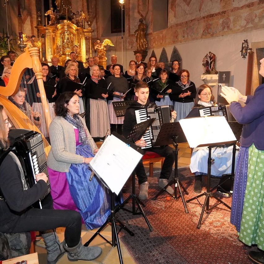 Ein Chor übt in einer Kirche mit Musikern, die Harfe, Akkordeon und andere Instrumente spielen. Ein Dirigent leitet, und die Chormitglieder lesen von ihren Notenblättern ab.