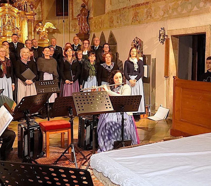 Eine Gruppe von Musikern tritt in einer Kirche auf, mit einer Flötenspielerin vorne, umgeben von Chormitgliedern. Die Musiker tragen formelle Kleidung.