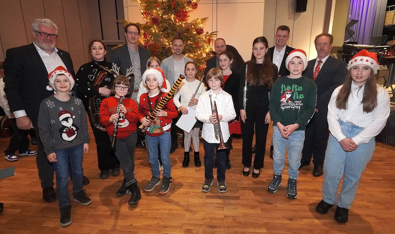 Eine Gruppe von Kindern und Erwachsenen posiert für ein Foto mit Musikinstrumenten in einer festlichen Umgebung. Die Kinder tragen Weihnachtsmützen und halten verschiedene Instrumente, während Erwachsene hinter ihnen stehen und lächeln. Ein Weihnachtsbaum ist im Hintergrund dekoriert.