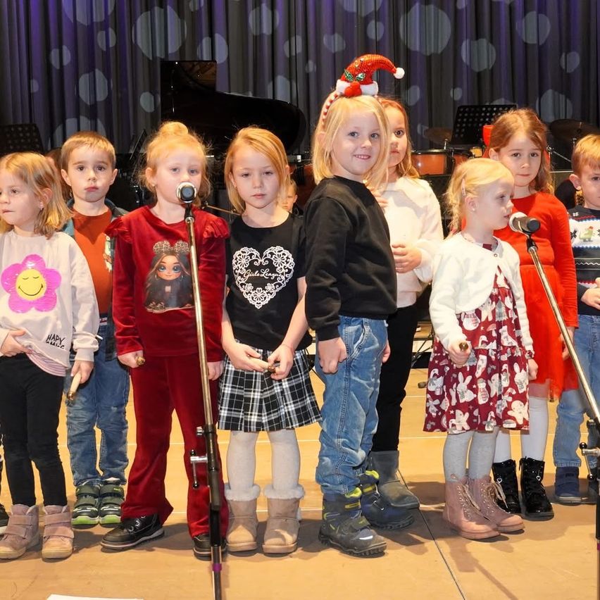 Eine Gruppe von Kindern, in Winterkleidung gekleidet, tritt mit Musikinstrumenten auf der Bühne auf. Hinter ihnen steht ein Klavier und Trommeln.