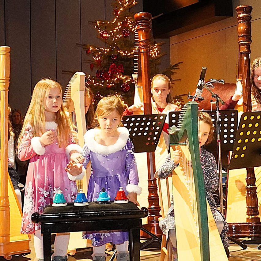 Eine Gruppe junger Mädchen in festlichen Outfits spielt Harfen und Glocken in einem dekorierten Raum, mit einem Weihnachtsbaum und Mikrofonen, die dahinter aufgestellt sind.