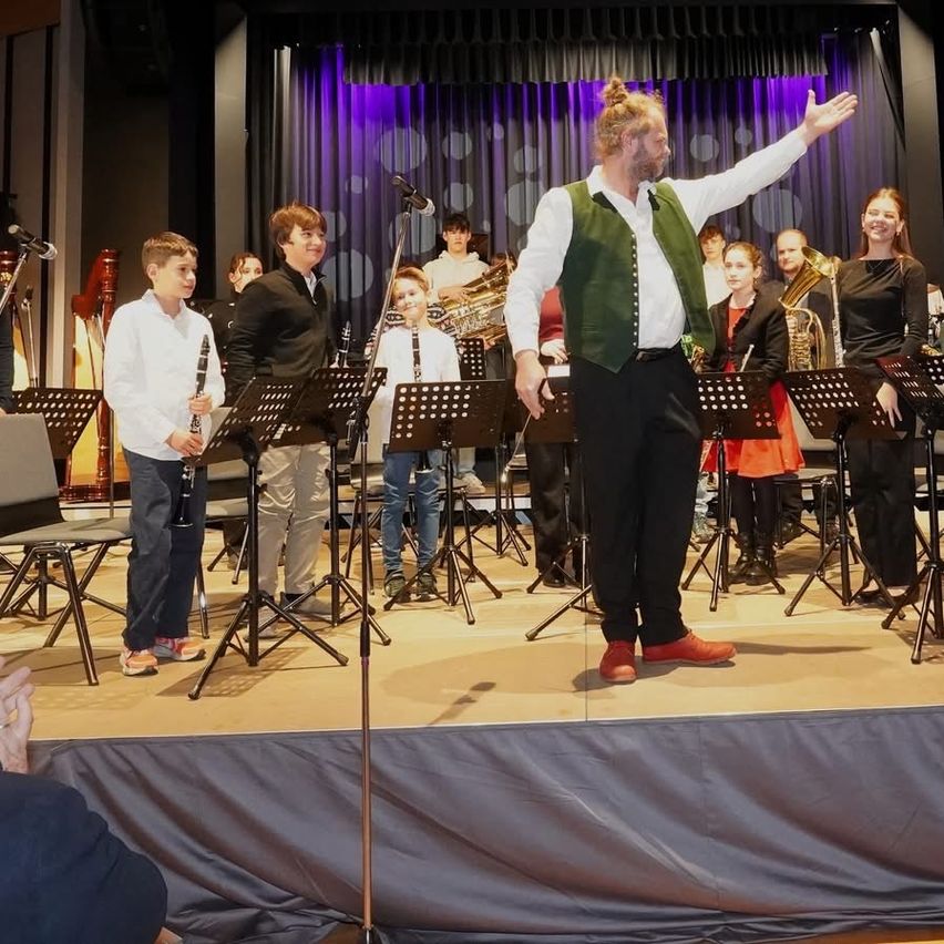 Eine Band spielt auf der Bühne mit verschiedenen Instrumenten. Ein Mann in der Mitte dirigiert und lächelt. Kinder umgeben ihn und spielen Instrumente wie Trompeten und Klarinetten.