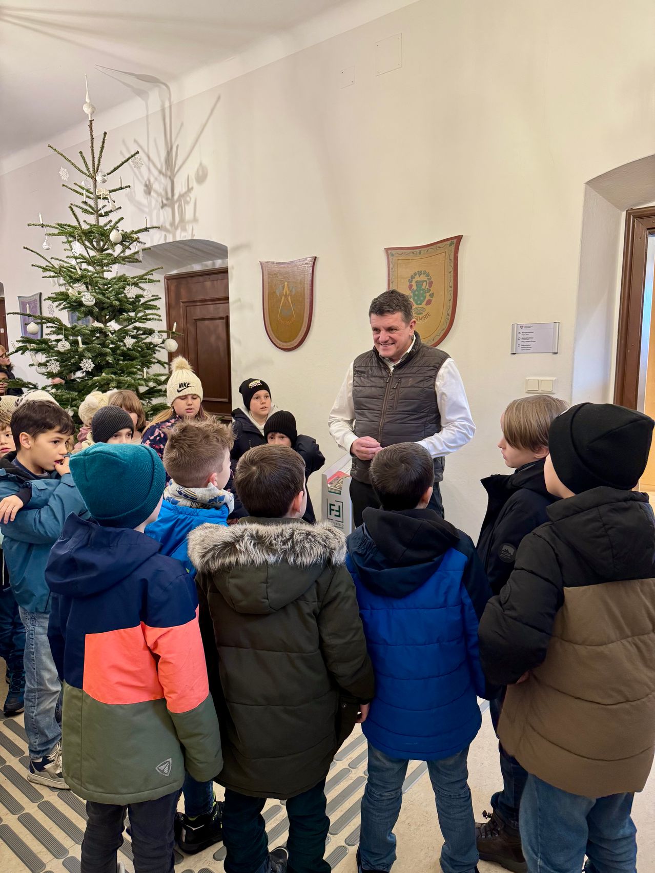 Eine Gruppe von Kindern steht um einen Mann in einer Weste herum, der sich neben einem Weihnachtsbaum in einem Raum mit weißen Wänden und einer Holztür befindet.