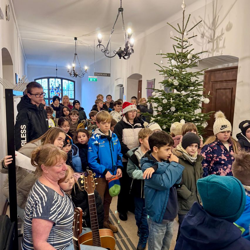 Eine Menschenmenge, einschließlich Kinder und Erwachsene, versammelt sich um einen Weihnachtsbaum in einem Flur. Eine Frau hält eine Gitarre, und einige Personen tragen Mützen und Brillen.