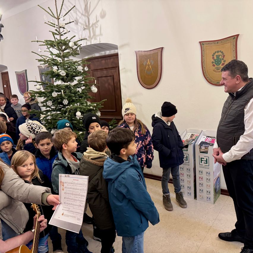 Eine Gruppe von Kindern steht um einen geschmückten Weihnachtsbaum herum, während ein Mann in einer Weste zu ihnen spricht. Der Raum hat eine weiße Wand, eine Holztür und zwei Banner. Einige Kinder halten Papiere, und eine Gitarre ist in der Nähe.