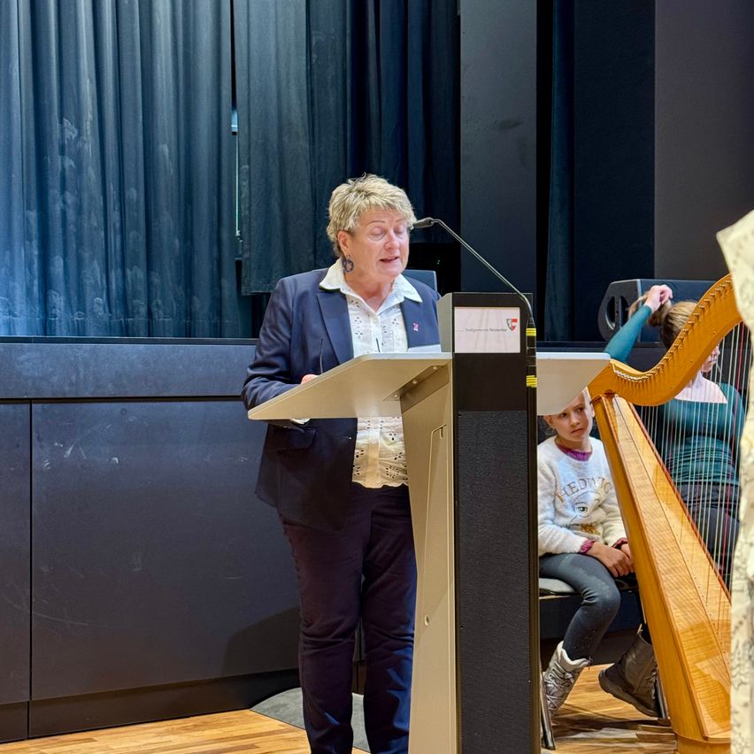 Eine Frau spricht an einem Podium mit Mikrofon, während ein junges Mädchen die Harfe spielt. Im Hintergrund befinden sich blaue Vorhänge und ein Holzboden.