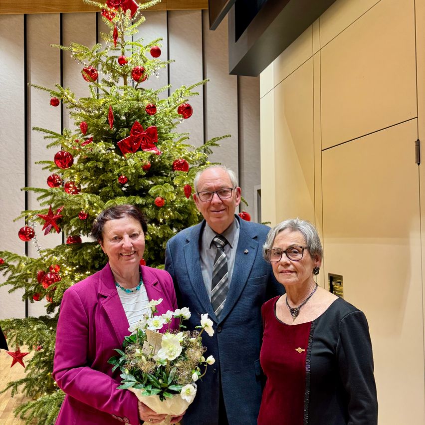 Drei Menschen stehen zusammen vor einem geschmückten Weihnachtsbaum und lächeln für ein Foto. Die Frau links hält einen Blumenstrauß.