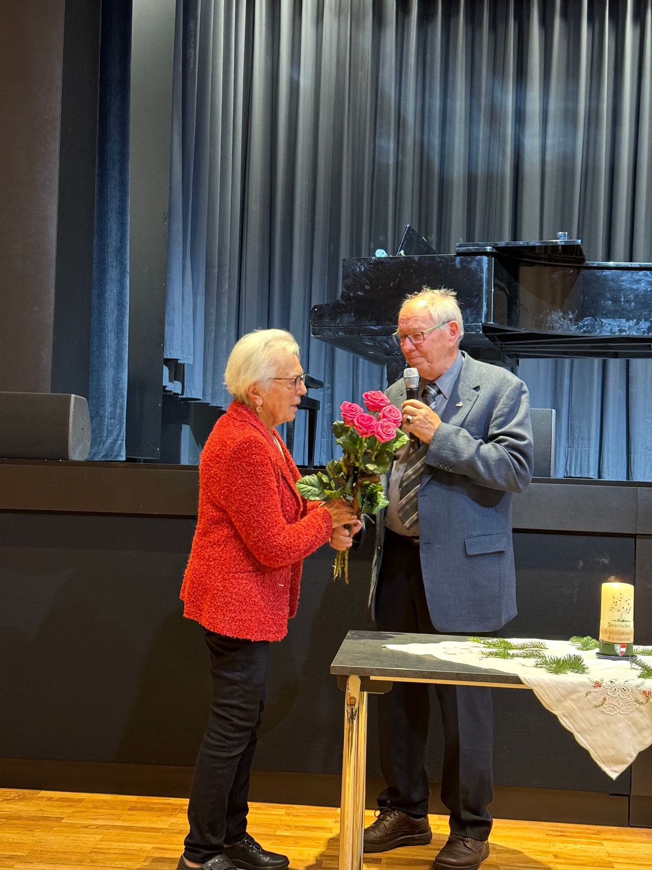 Eine ältere Frau und ein Mann stehen gemeinsam vor einer Bühne mit einem Klavier, der Mann hält einen Strauß roter Rosen und ein Mikrofon, während die Frau den Strauß hält.