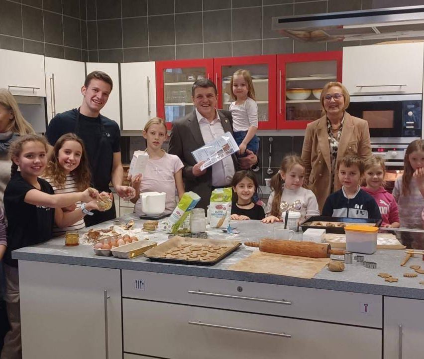 Eine Gruppe von Menschen in einer Küche mit einem großen Tresen. Sie lächeln und scheinen Kekse zuzubereiten. Keksablagen und Zutaten stehen auf dem Tresen.