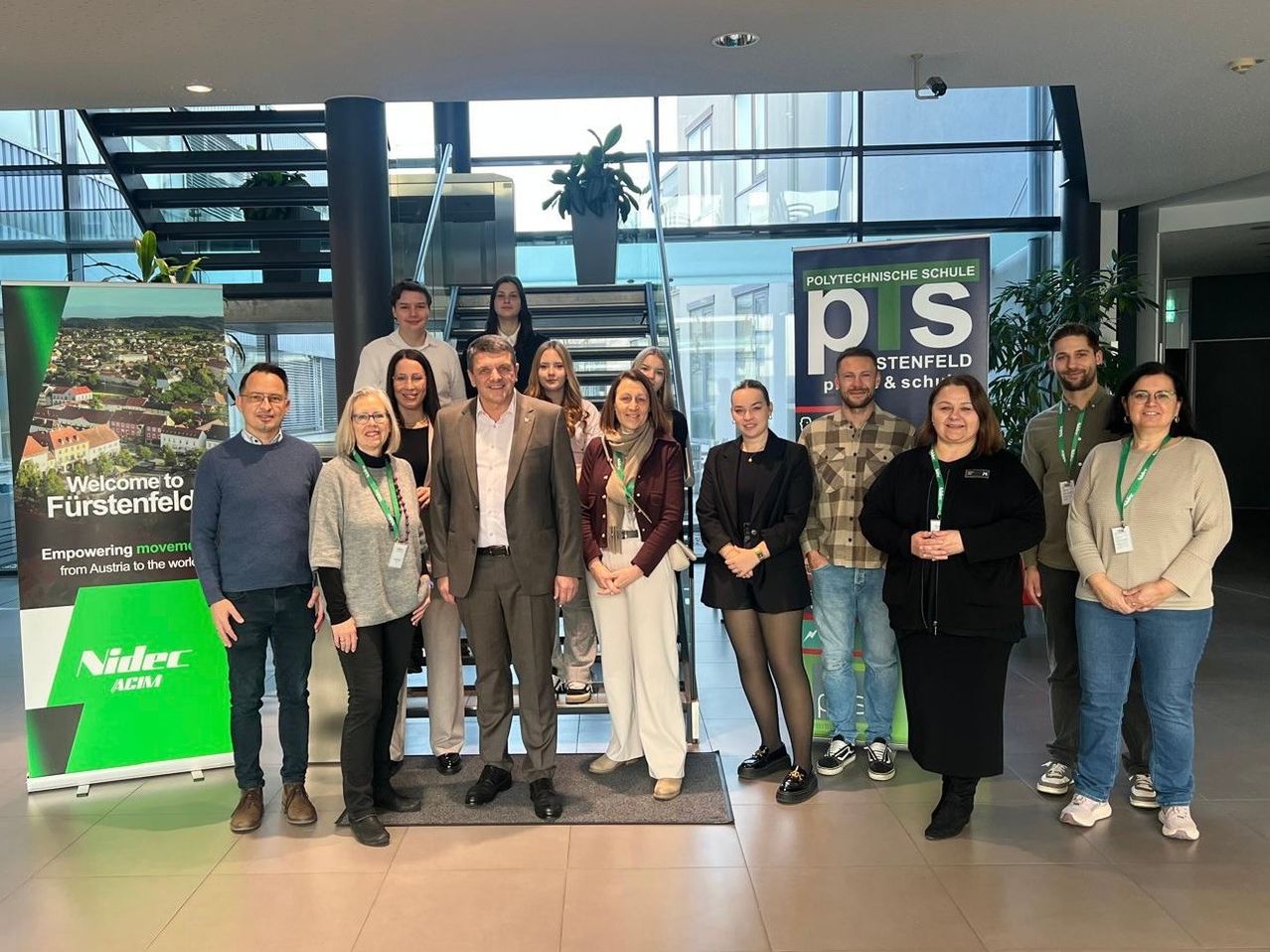 Eine Gruppe von Menschen steht drinnen, wahrscheinlich in einer Schule, und trägt Ausweise. Dahinter befindet sich eine Treppe mit einem grünen Schild. Ein Mann im Anzug lächelt und hält ein Schild.