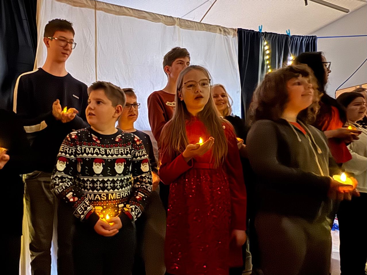 Eine Gruppe von Kindern und Erwachsenen in einem Zelt, die Kerzen halten, einige mit Brillen und Weihnachtspullovern, möglicherweise bei einer Weihnachtsveranstaltung.