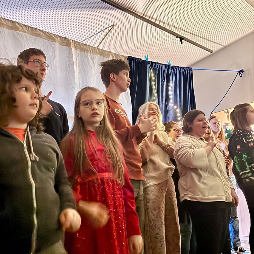 Eine Gruppe von Menschen, darunter Kinder und Erwachsene, steht in einer Reihe, wahrscheinlich in einer Kirche, und nimmt an einem Chorauftritt teil.