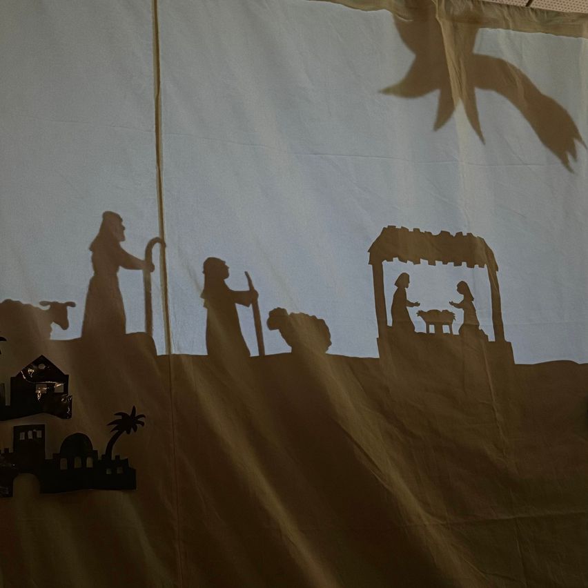 Eine Krippe wird auf einem hellen Hintergrund dargestellt, mit einer Silhouette einer Person und eines Kindes in der Nähe einer Krippe. Der Hintergrund zeigt eine Silhouette einer Person mit einem Stab.