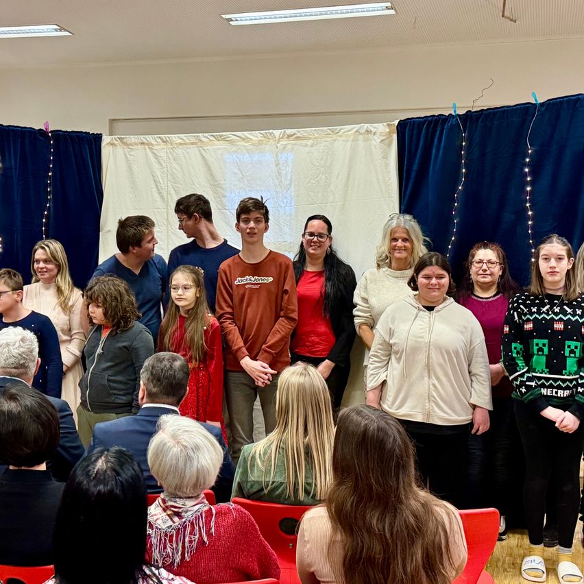 Eine Gruppe von Menschen steht vor einer Bühne mit blauen Vorhängen, lächelt und schaut in die Kamera. Ein Junge in einem roten Shirt steht in der Mitte. Ein Mann und eine Frau sitzen in der vorderen Reihe.