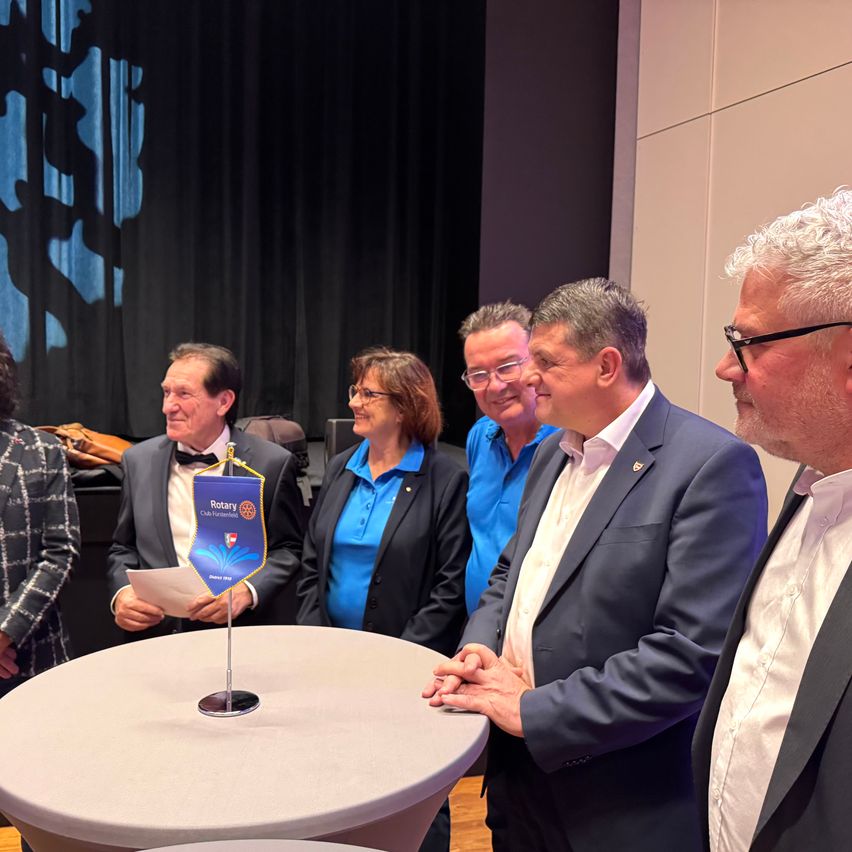 Eine Gruppe von Menschen steht um einen Tisch mit einer Rotary-Fahne, alle formell gekleidet. Ein Mann auf der rechten Seite trägt eine Brille. Sie scheinen zu sprechen und zu lachen.