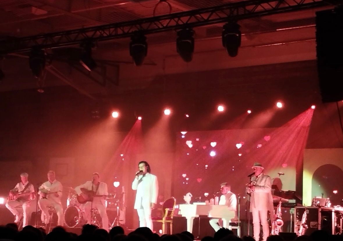 Eine Band tritt auf der Bühne mit roten Lichtern und herzförmigen Projektionen auf. Ein Mann singt, während andere Instrumente spielen.